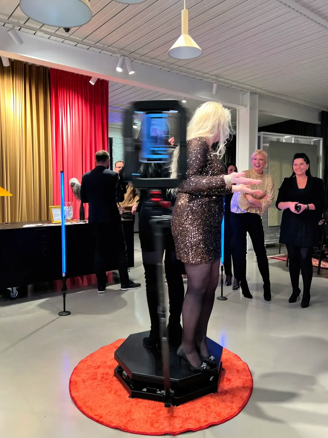 Viese spīdīgā kleitā dejo uz 360 video spinera platformas uz sarkana paklāja, apkārt vērojoši viesi