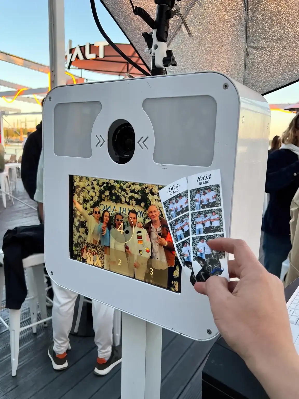 Fotoautomāts Rīgā: viesis tur tikko izdrukātu foto loksnīti pie ekrāna ar grupas bildi