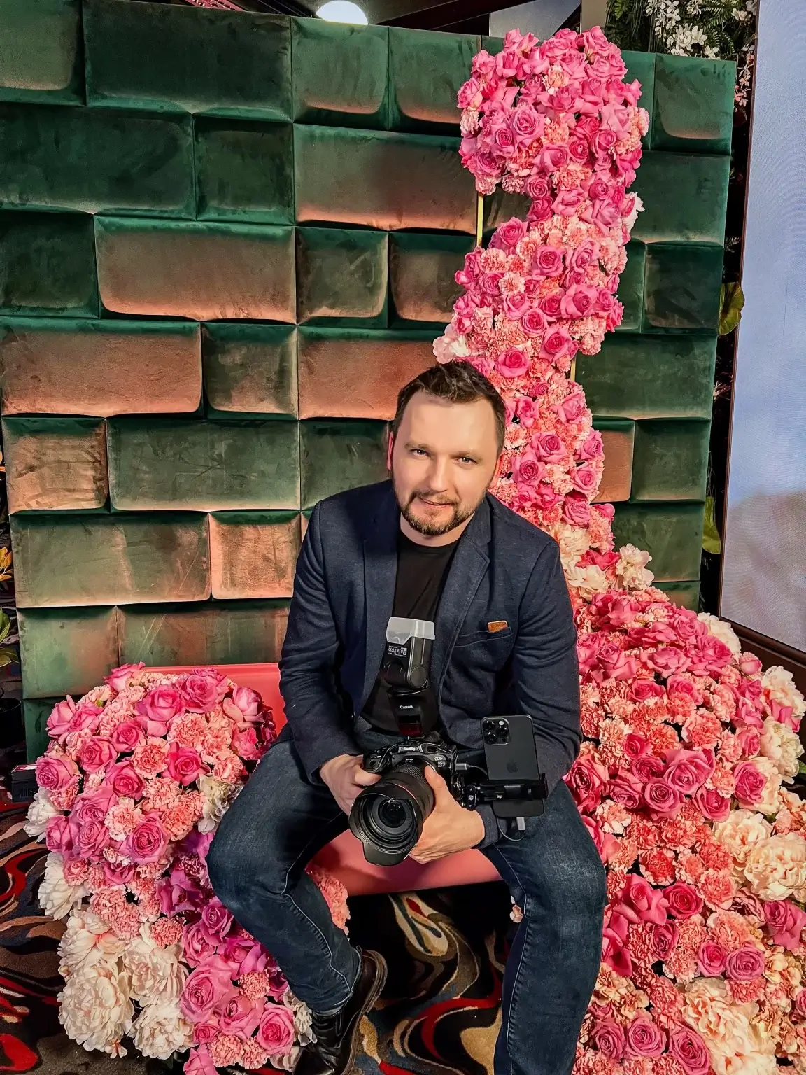 Fotogrāfs ar kameru sēž rozā ziedu dekorā pie zaļas ķieģeļu sienas